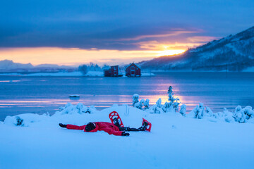 A man with snowshoes under moody sky winter sunset blue hour over the red houses in Norway