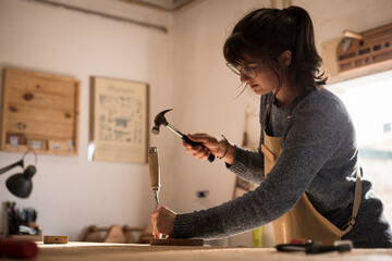 A young female carpenter working as wood designer in a small carpentry workshop. Young business woman handcrafting a piece of timber and designing new house furniture. Entrepreneurs concept lifestyle - obrazy, fototapety, plakaty