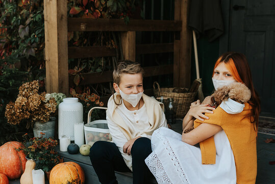 children a boy and a girl with their poodle dog in protective masks on the porch of the backyard decorated with pumpkins in autumn, the concept of the covid-19 pandemic quarantine