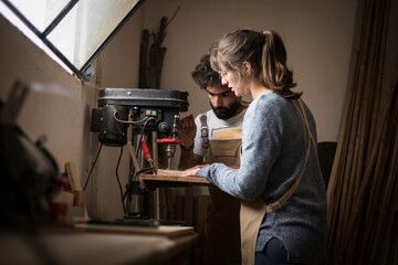 A young couple of carpenter working together in a small wood laboratory using a drill machine for timber. Couple crafting new home furniture in a carpentry workshop. young entrepreneurs concept