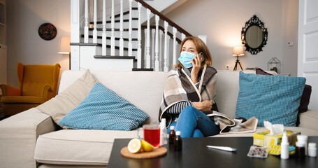 Sick woman sitting on sofa at home in medical face mask and calling with cell telephone. Cold and fever concept. Coronavirus.