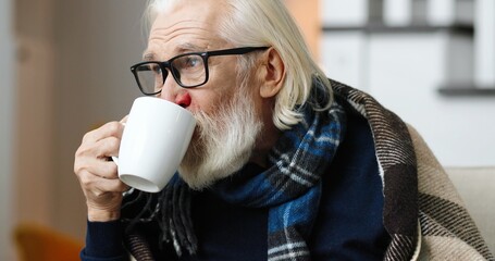 Close up portrait of sick senior man sitting on sofa at home drinking hot tea.