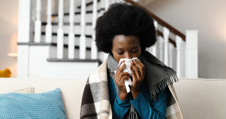 Sick African American woman wrapped blanket blowing running nose sitting on sofa at home. Flu...