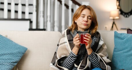 Portrait of beautiful sad sick Caucasian woman in plaid sitting on couch at home with cup of hot drink. Female not feeling well and having virus infection. Woman with cup of tea. Treatment concept