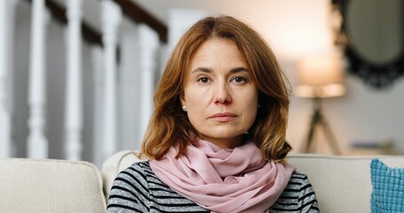 Close up portrait of beautiful Caucasian woman with scarf sitting on sofa in living room and looking at camera. Pretty sick female not feeling well at home indoor. Health concept. Virus infection