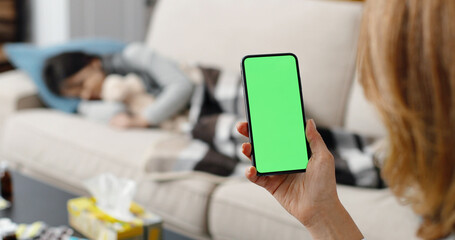 Caucasian female holding smartphone with green screen in hand while sitting at home. Woman holds cellphone with chroma key and looking at display. Sick girl laying on sofa. Indoors concept