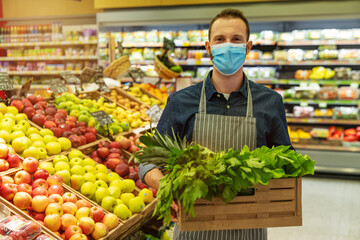Authentic shot of male staff worker wearing medical mask is carrying a wooden crate with the fresh vegetables is looking in camera satisfied with her work in organic section at supermarket.