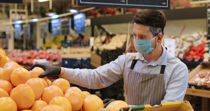 Portrait Of Male Food Store Employee In Face Protection Shield And Apron Standing In Store And Sorting Fruits. Handsome Caucasian Man Seller In Mask Working Indoor. Supermarket Concept