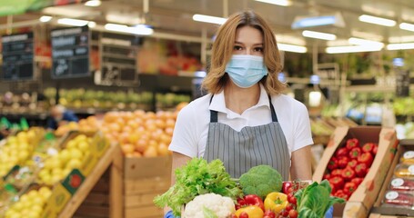 Camera approaching female seller in mask and apron standing in supermarket with packet with food. Close up of beautiful Caucasian woman worker with vegetables in grocery store alone. Retail concept