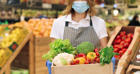 Camera approaching female seller in mask and apron standing in supermarket with packet with food. Close up of beautiful Caucasian woman worker with vegetables in grocery store alone. Retail concept