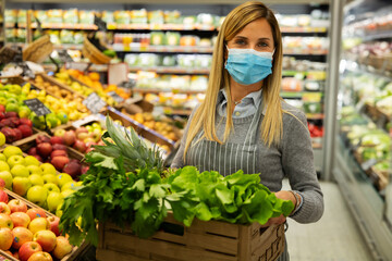 Authentic shot of female staff worker wearing medical mask is carrying a wooden crate with the fresh vegetables is looking in camera satisfied with her work in organic section at supermarket