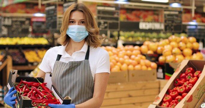 Portrait of beautiful Caucasian woman in face mask and apron standing in food store with red hot chili papers indoors. Pretty female seller holding box with food in supermarket. Retail concept