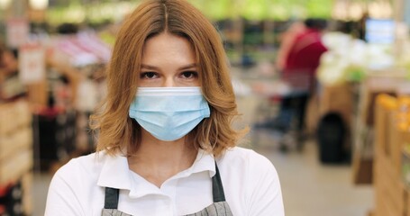 Close up portrait of beautiful female worker in medical mask and apron looking at camera while...