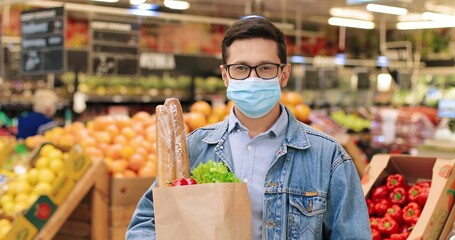 Close up portrait of joyful handsome male standing in grocery store with packet with food. Camra approaching Caucasian man in mask with vegetables in supermarket in quarantine. Food concept