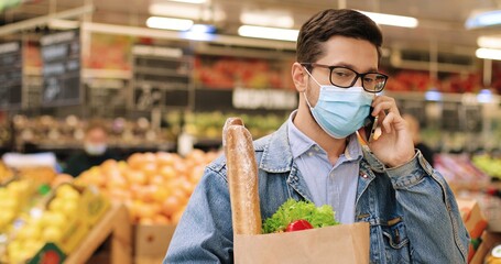 Close up portrait of handsome Caucasian male standing in grocery store with packet with food and talking on smartphone. Man in mask and glasses with vegetables in supermarket calling on cellphone