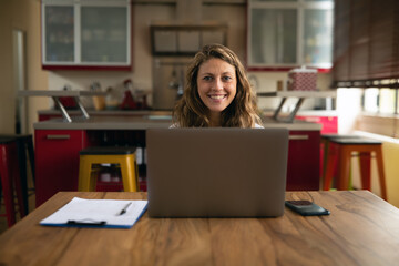 Fototapeta na wymiar Authentic shot of young successful businesswoman is smiling satisfied with her work in camera while doing smart working with laptop from home in the kitchen during lockdown. 