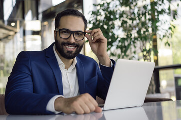 Young confident businessman using laptop computer, looking at camera, smiling. Portrait of handsome...