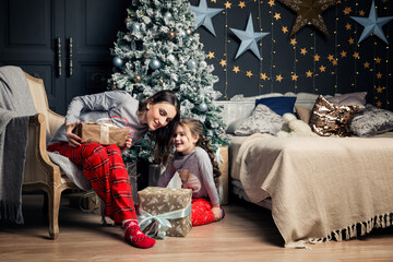 Obraz premium Mom and daughter in pajamas unpack New Year's gifts under the Christmas tree. Mother kisses her daughter