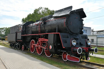 Plakat Close up on a vintage train with a black metal body and red and white wheels standing on a side track surrounded with some trees and with a train station visible in the background in Poland