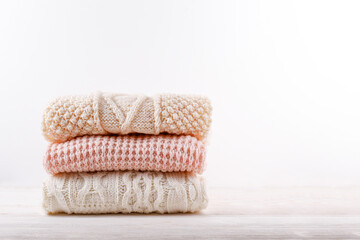 Fototapeta na wymiar Bunch of knitted warm pastel color sweaters with different knitting patterns folded in stack on white wooden table, white wall background. Fall winter season knitwear. Close up, copy space for text