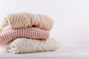 Fototapeta na wymiar Bunch of knitted warm pastel color sweaters with different knitting patterns folded in stack on white wooden table, white wall background. Fall winter season knitwear. Close up, copy space for text