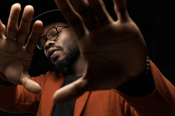 portrait photo of a black man with glasses, black hat, brown jacket and jacket on a dark background in the studio, showing his palms to the camera - 385750255