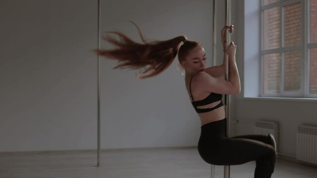 Slim young female in high heeled shoes dancing on floor and near pole in slow motion during performance in modern studio