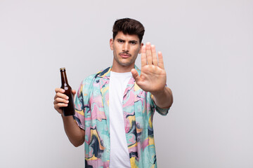young man with a beer looking serious, stern, displeased and angry showing open palm making stop...