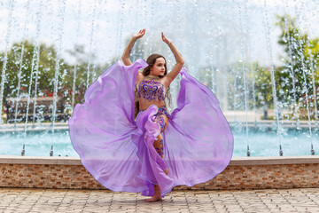 pretty caucasian girl in traditional belly dancer costume of lavender color. dancing girl. Fountain on the background
