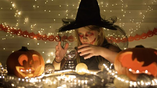 Teenage girl dressed as a witch with scary makeup on her face. Girl conjures over a magic ball. Dark gold background. Halloween.