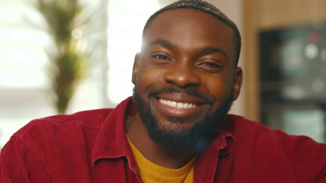 Face young handsome african american man looking at camera smiling sit on sofa at home. Relax. Attractive guy. Close up. Slow motion
