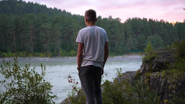 A teenager stands on the riverbank on a cliff. He looks at the mountains