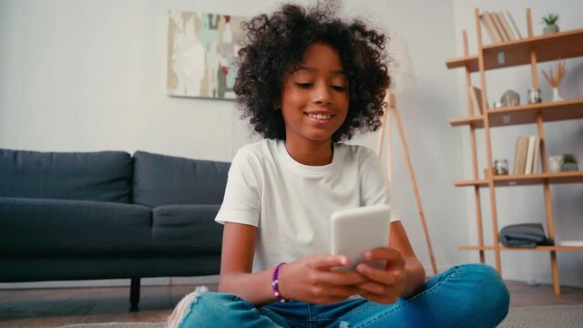 African american girl sitting on floor and texting on cellphone at home