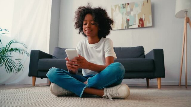 African american girl with crossed legs using cellphone while sitting on carpet