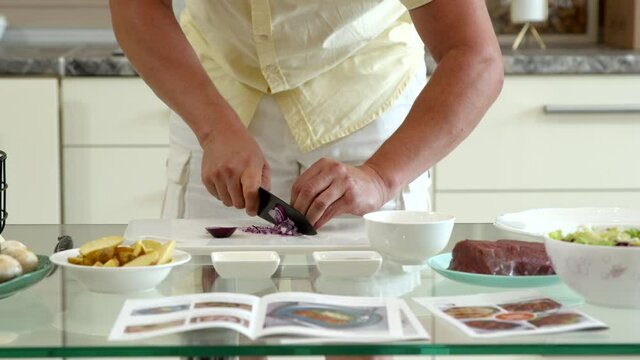man slice onion, looking at cookbook recipe at home. All ingredients are neatly arranged on table. Novice chef learns to cook. man in kitchen cooks culinary surprise for his wife. Mother's Day Concept