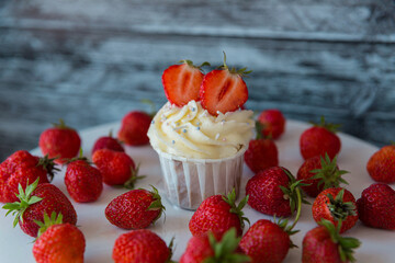 cupcake in the center with white cream decorated with ripe strawberries on a gray blue background and scattered strawberries.
