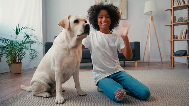 African american girl stroking dog and waving at camera while sitting on carpet