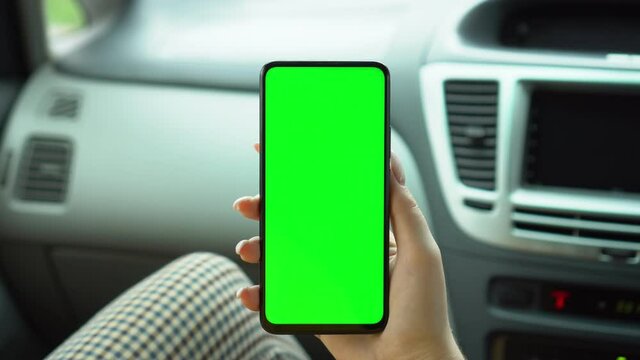 A hand holding a smartphone with green blank screen in the car for direction, massage, location, business. Woman sits in modern car and works on smartphone - green screen - closeup. Chroma key.