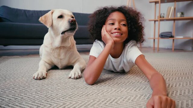 African American Girl Waving Hand Near Dog Lying On Carpet At Home