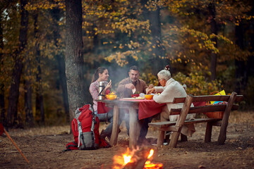 An elderly couple and their daughter enjoying the food at picnic in the forest around a campfire