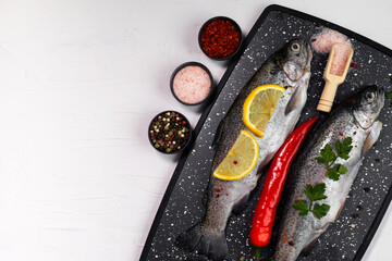 Fresh rainbow trouts on black board on table with salt, pepper, lemon and chili pepper ingredients, flatlay, copy space