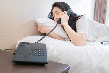 Young sleeping girl trying to pick up the phone in bed room while sleeping. Women lazily pick up the room telephone while lying on bed feeling sleepy. Young girl in white on bed in morning.