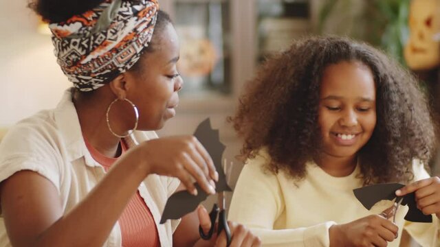 Cute Afro-American girl smiling and chatting with mother while making paper bats for Halloween decorations at home