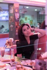 Young beautiful retro girl sitting in a diner restaurant. Woman in black eating in a diner with beautiful neon light. Pretty happy young woman sitting in the diner. Fashion girl in a vintage cafe.
