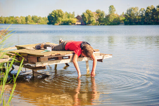 a young boy lies on a fishing bridge with a toy boat and dangles his arms into the water of the lake