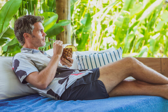 Middle age caucasian man sitting on a couch relaxing alone drinking a pineapple cocktail indoors. Male enjoining his vacation isolated during covid-19 pandemic.