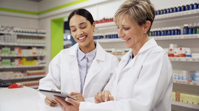 Two female colleagues scrolling through emails on digital tablet standing behind prescription counter in pharmacy 