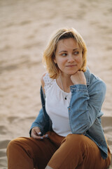 Fototapeta na wymiar beautiful young caucasian smiling woman wearing white blouse and denim jacket stitting on sand at the beach before sunset, touching hair and looking aside. Image with selective focus