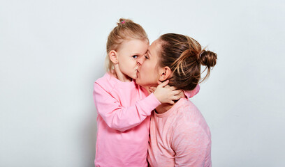 Little daughter hugging and kissing her mother spending time together in pink clothes over gray...