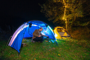 The guy prepares food, a mini gas burner. A pot and a bottle of water. Overnight in a tent. Camping, A man sits inside. The light of the gerlands. Night. Leisure and hobbies. Hiking. Copy space